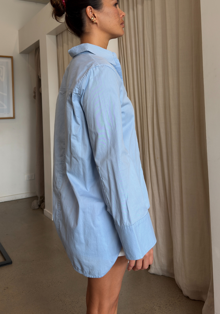 A woman wearing an oversized light blue button-up shirt, shown from the side in a home interior with beige curtains and neutral walls.