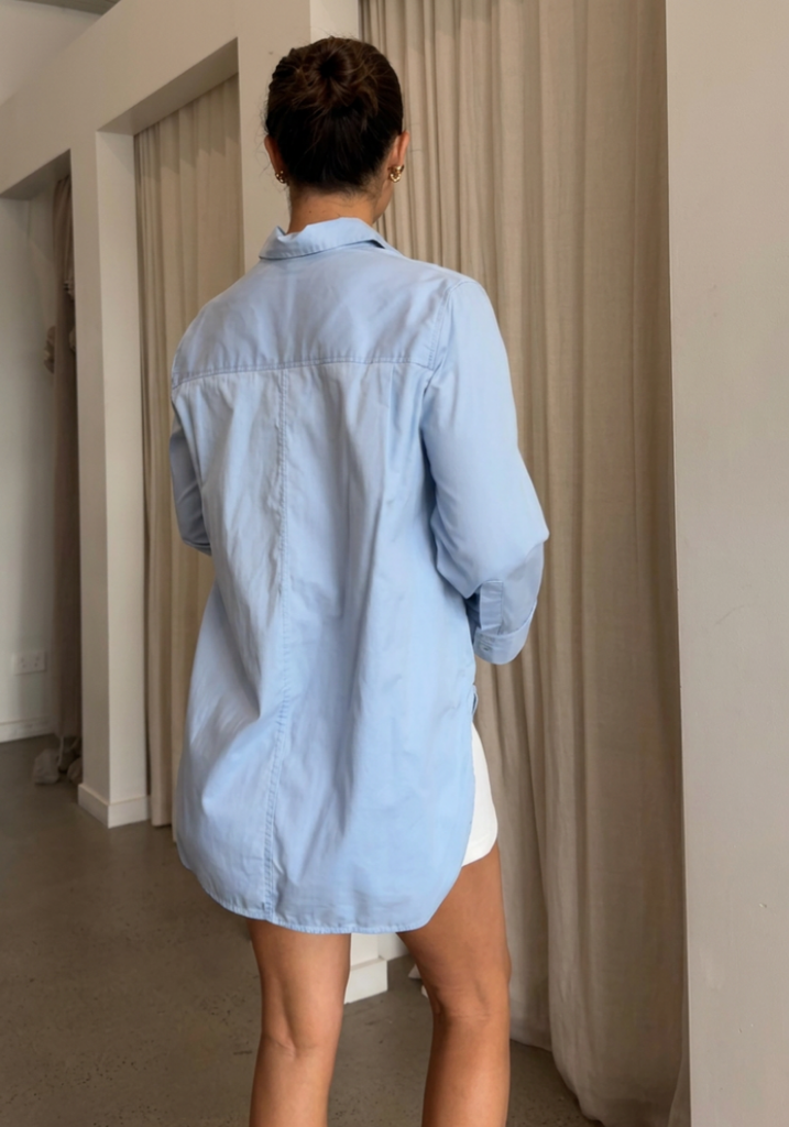 A woman viewed from behind wearing an oversized light blue button-up shirt and white shorts in an indoor room with beige curtains.